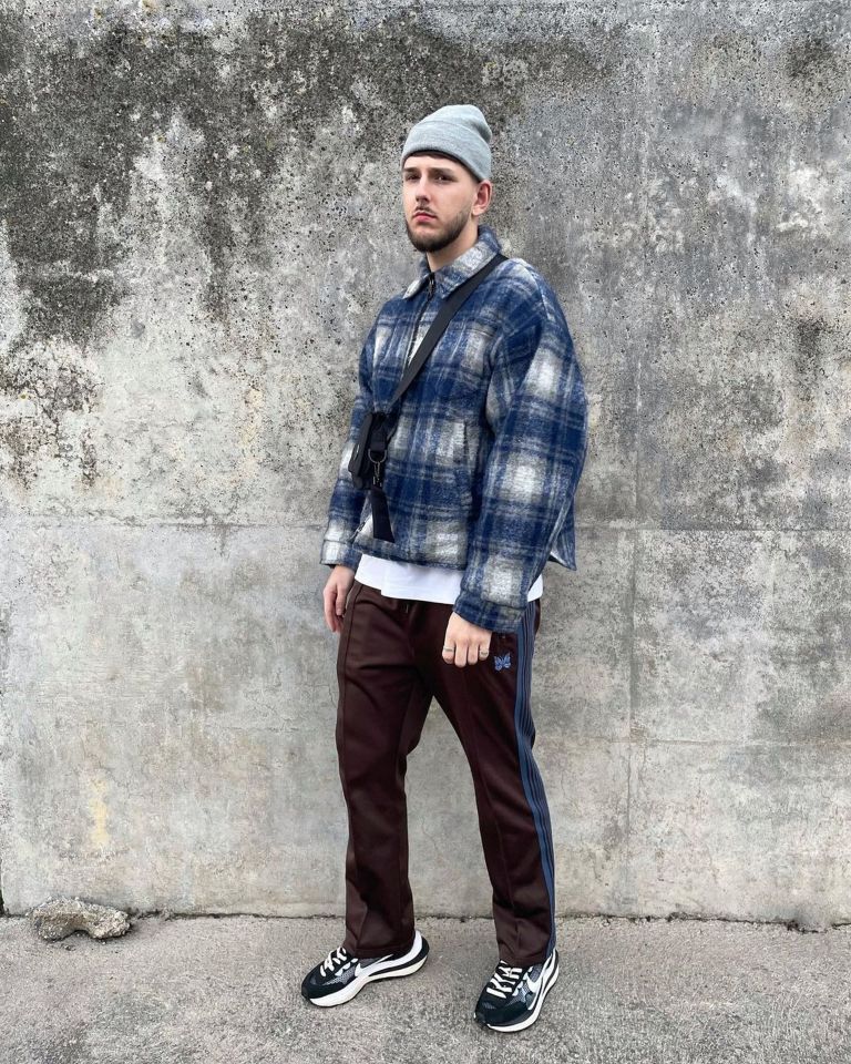 a man wearing a plaid jacket and brown pants is standing in front of a concrete wall