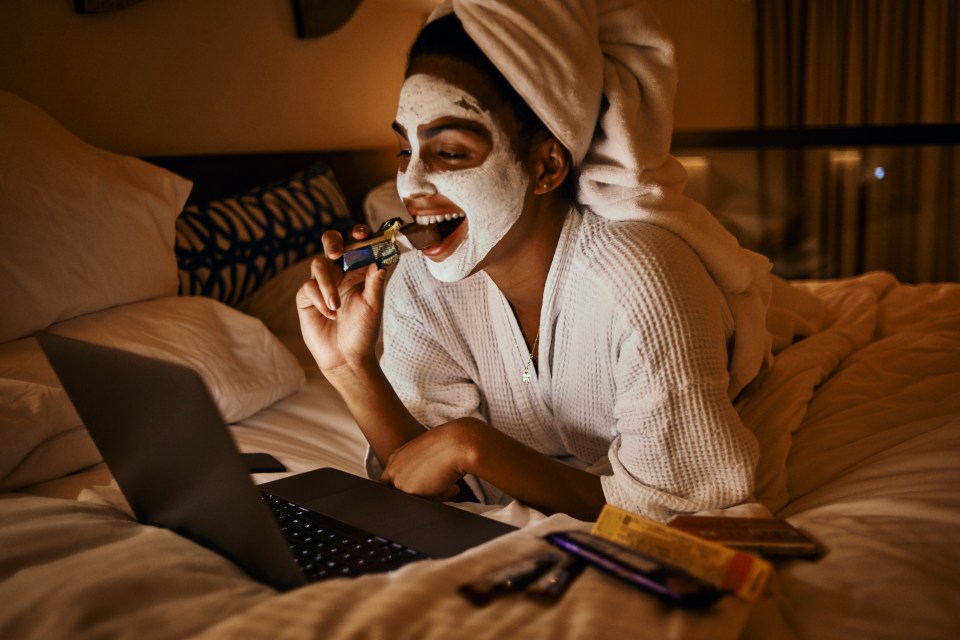 a woman with a mask on her face is eating a chocolate bar while looking at a laptop