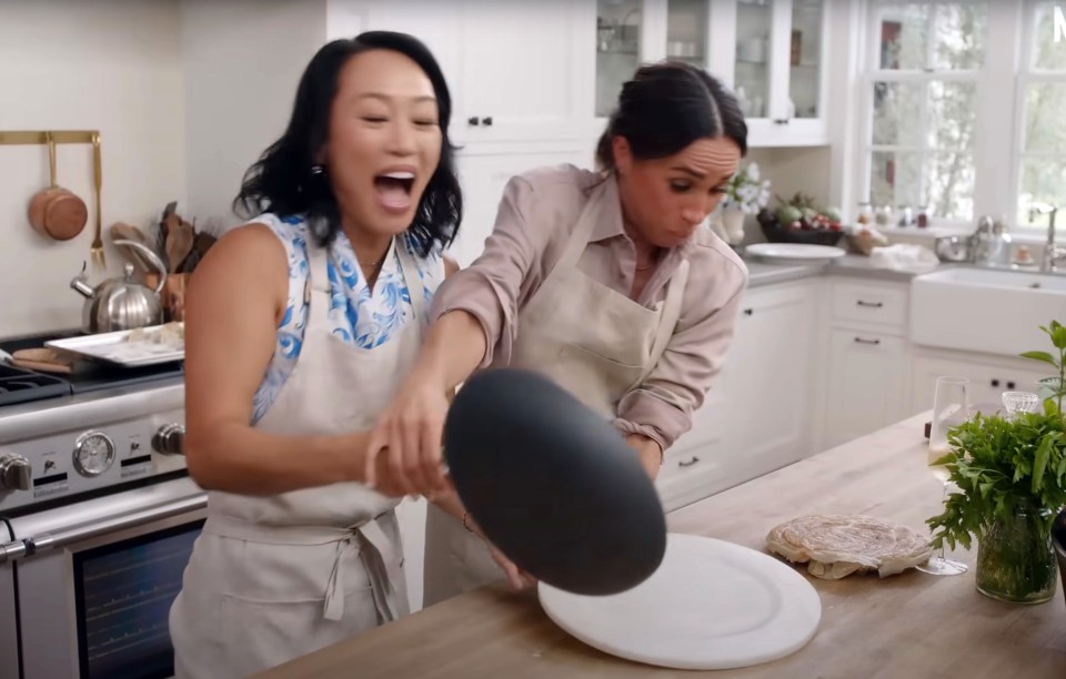 Meghan Markle and a friend laughing while cooking in a kitchen.
