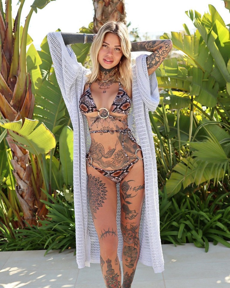 Woman with extensive tattoos wearing a snakeskin-print bikini and light gray cardigan.