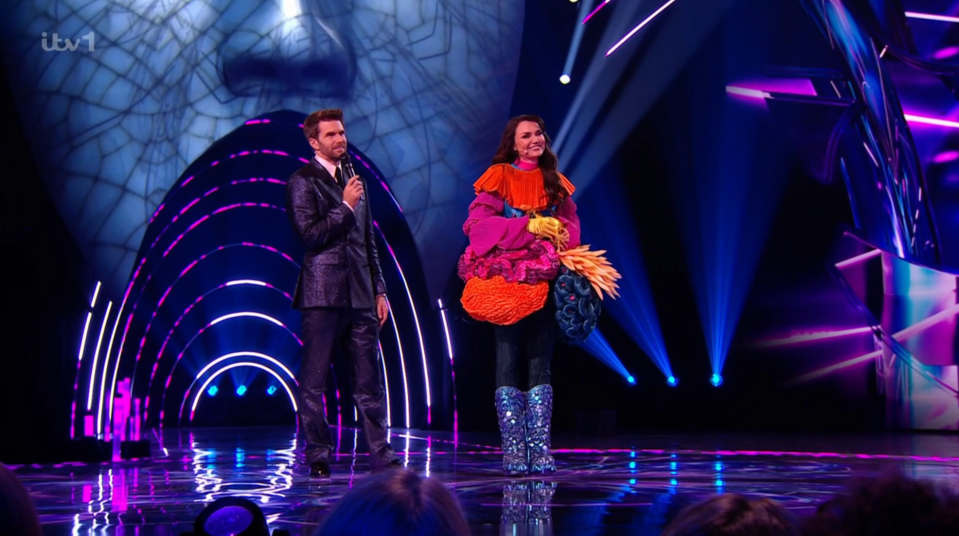 Screenshot of a man and woman on a stage; the woman is wearing a large, colorful costume.