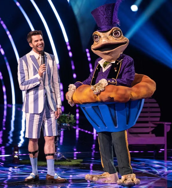 A man in a striped suit interviews a contestant dressed as a frog in a cupcake costume on The Masked Singer.