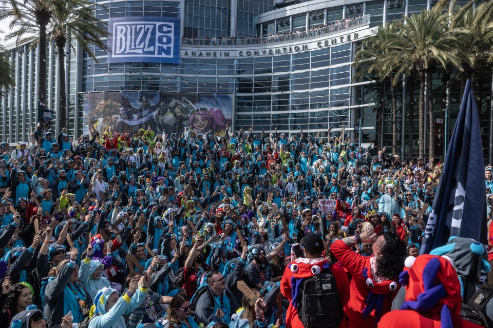Large group of people in blue costumes at BlizzCon, holding up wooden swords.