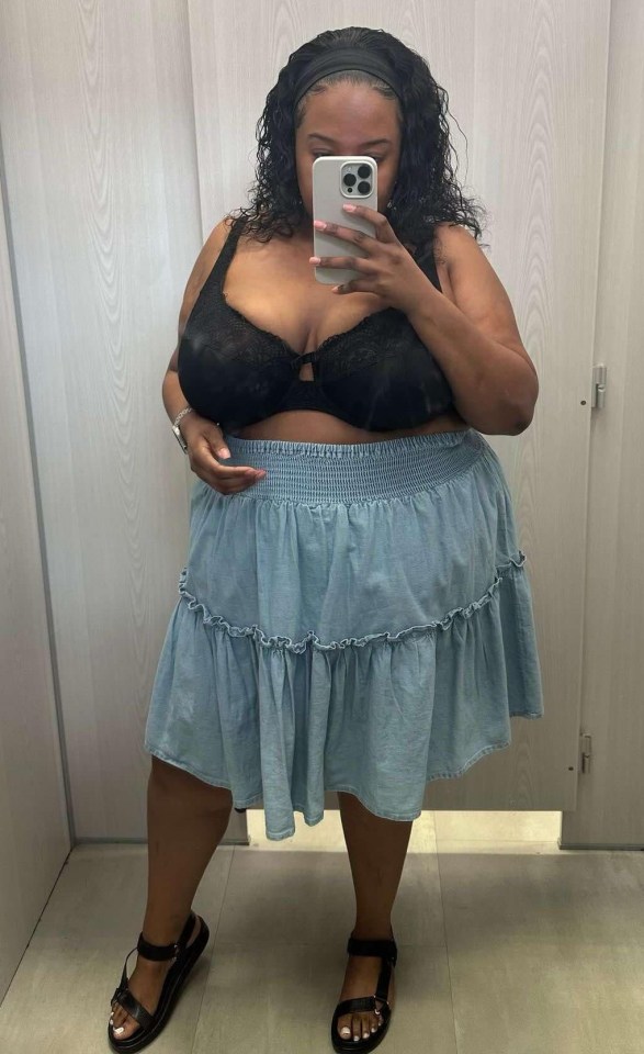 Woman taking a selfie in a changing room wearing a black bra and a light blue tiered skirt.