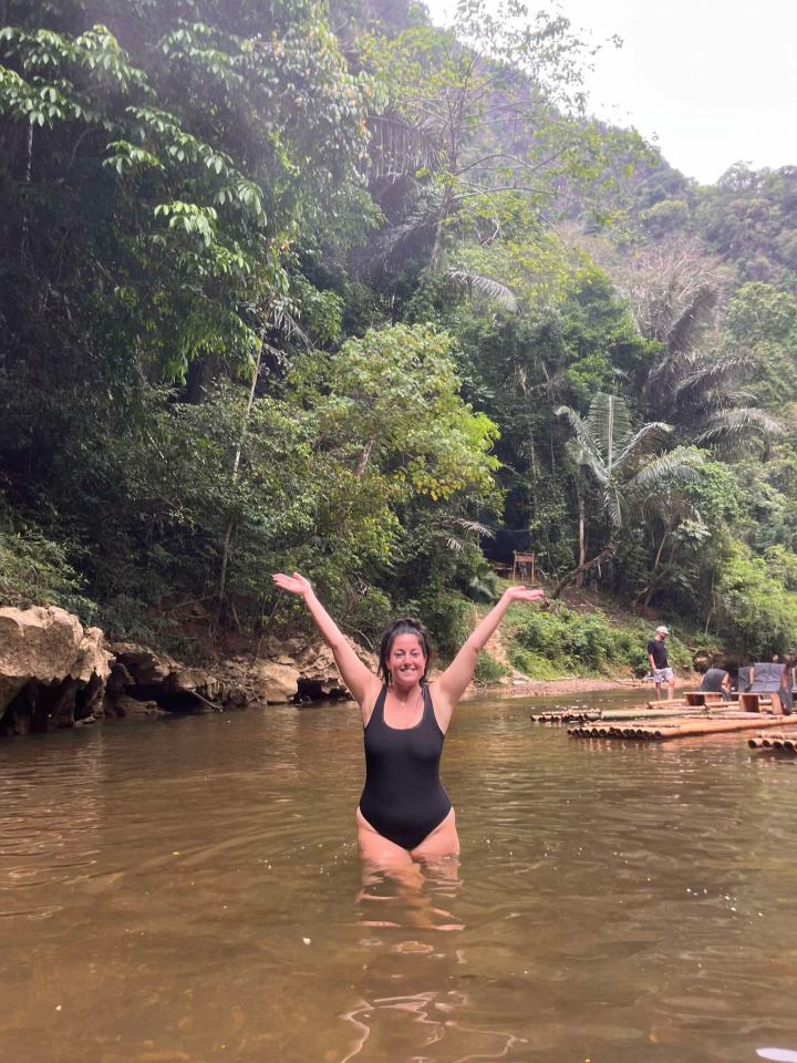 Woman in a black swimsuit wading in a river, arms outstretched.  Bamboo rafts are visible in the background.