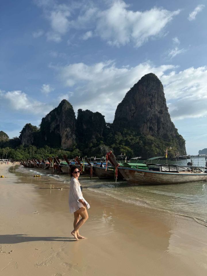 Woman on a beach in Thailand with longtail boats and limestone cliffs in the background.