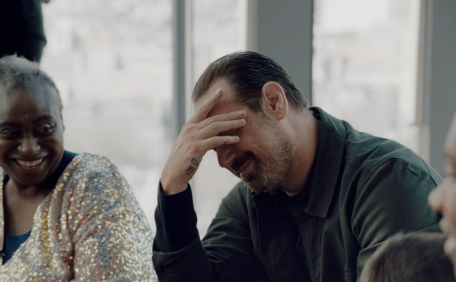 Man laughing with hand on forehead, woman smiling in background.