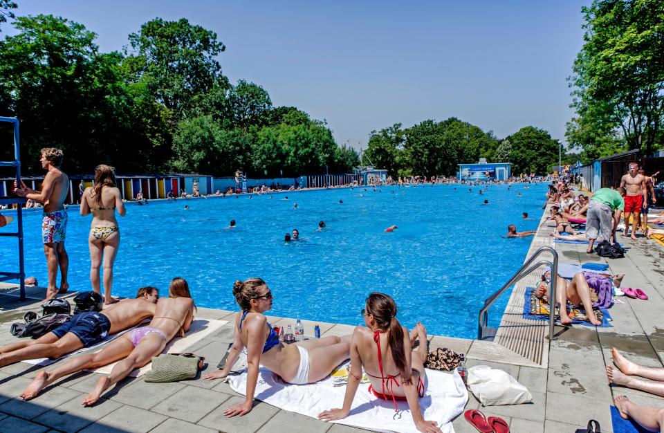 Crowded outdoor swimming pool with people swimming and sunbathing.