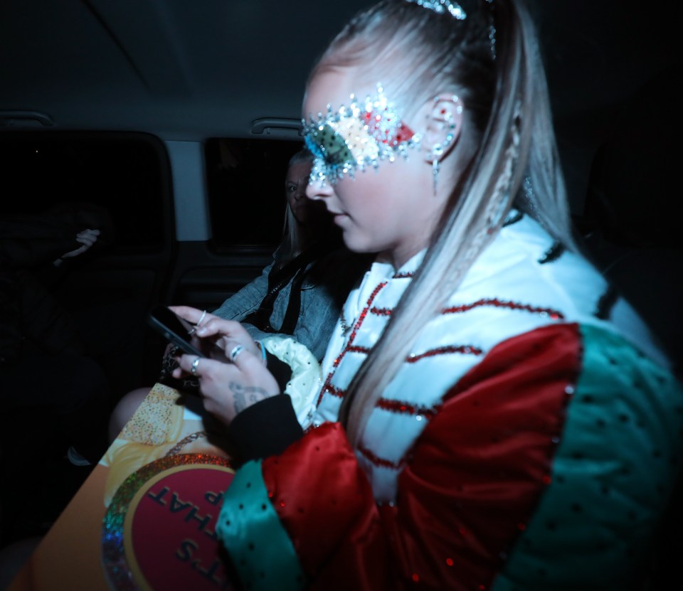 Woman wearing a sparkly mask and red and teal jacket uses a smartphone in a car.