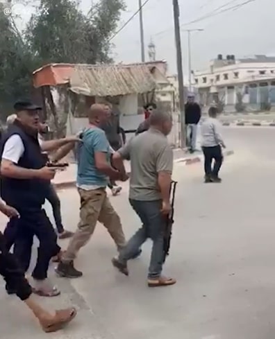 A group of armed men escorting a man in a light blue shirt down a street.