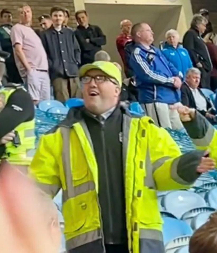 A man in a bright yellow safety jacket leads a crowd in a stadium.