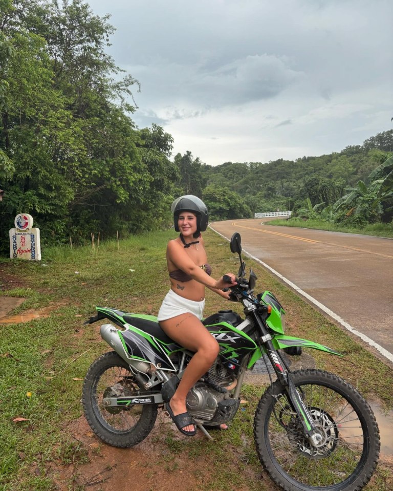 A young woman sits on a green motorcycle on a road in Thailand.