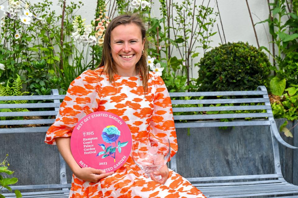 Woman in orange dress holding a gold medal and glass award in a garden.