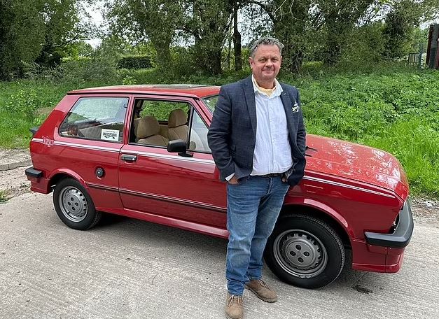 Man standing beside his red 1980s car.
