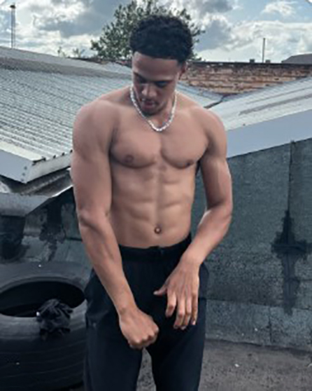 Shirtless man with defined abs, wearing black pants and a silver necklace, standing outdoors.